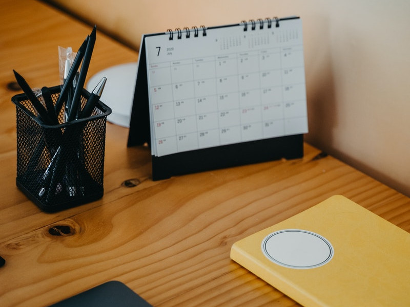 Imagen de una mesa de escritorio sobre la que se encuentra una agenda de color amarillo, un portalápices con varios elementos de escritura y un calendario, en referencia al Día Internacional de la Tecnología Social promovido por FUNTESO.